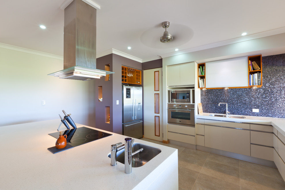 Bild på ett funkis l-kök, med en dubbel diskho, släta luckor, vita skåp, blått stänkskydd, stänkskydd i mosaik, rostfria vitvaror och en köksö