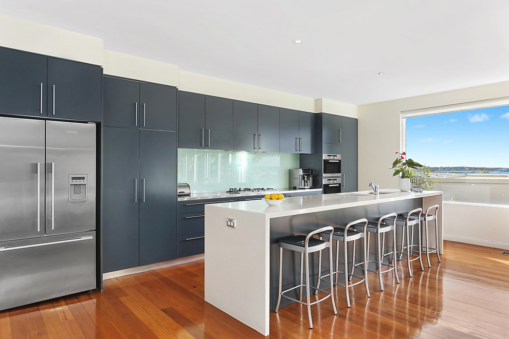 Cette image montre une cuisine parallèle design avec un placard à porte plane, des portes de placard grises, une crédence bleue, un électroménager en acier inoxydable, un sol en bois brun, îlot, un sol marron et un plan de travail blanc.