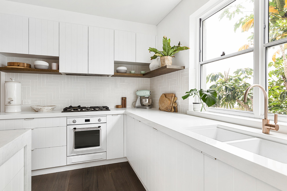 Example of a mid-sized beach style l-shaped dark wood floor and brown floor kitchen pantry design in Sydney with an undermount sink, shaker cabinets, white cabinets, quartz countertops, beige backsplash, ceramic backsplash, white appliances, an island and white countertops