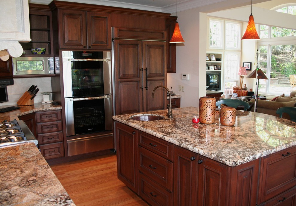 Example of a large classic l-shaped medium tone wood floor open concept kitchen design in San Francisco with an undermount sink, raised-panel cabinets, dark wood cabinets, granite countertops, beige backsplash, stone tile backsplash, stainless steel appliances and an island