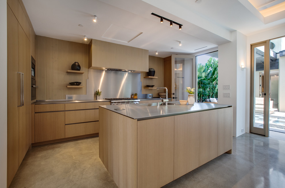 Photo of a beach style kitchen in Los Angeles with an integrated sink, flat-panel cabinets, light wood cabinets, stainless steel worktops, metallic splashback, stainless steel appliances, concrete flooring, an island and grey floors.