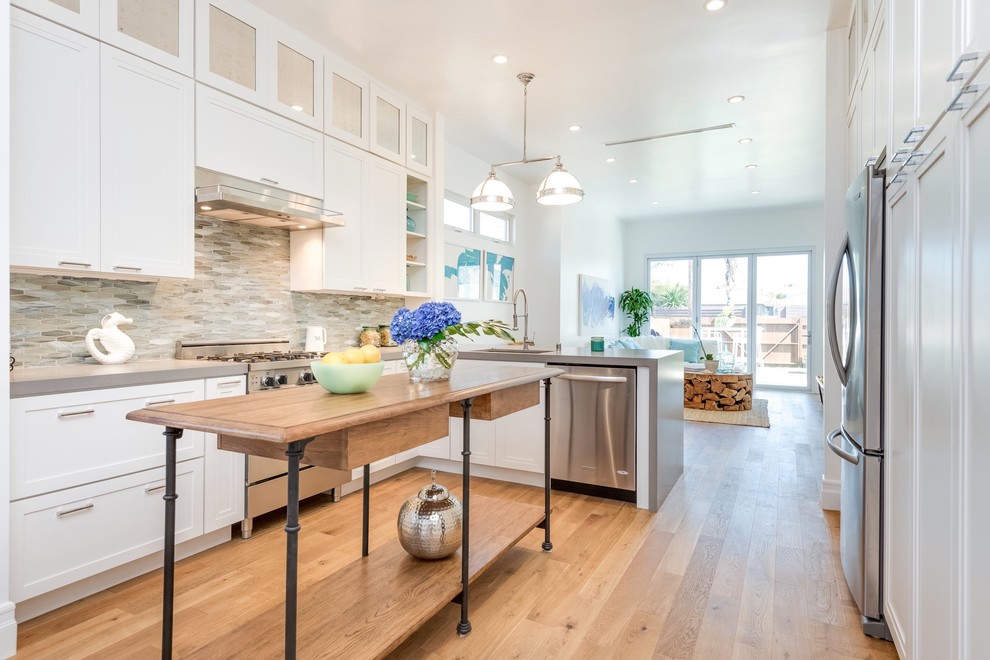 Maritime Wohnküche in U-Form mit Schrankfronten mit vertiefter Füllung, weißen Schränken, Kücheninsel, Küchenrückwand in Grau und braunem Holzboden in Los Angeles