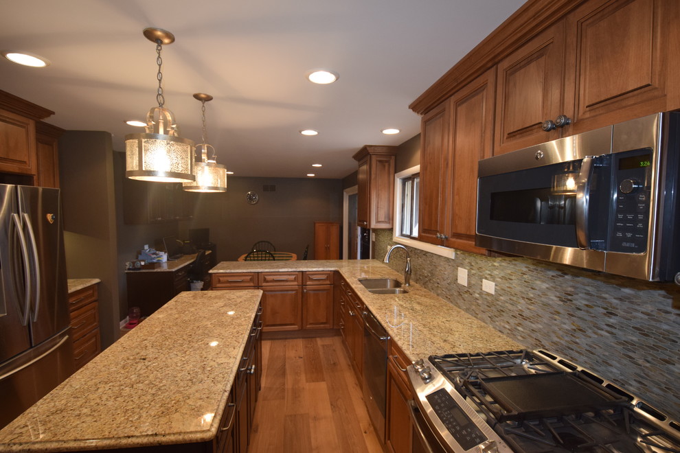 This is an example of a traditional kitchen in Chicago with raised-panel cabinets, brown cabinets, granite worktops, grey splashback, glass tiled splashback, stainless steel appliances, medium hardwood flooring and an island.