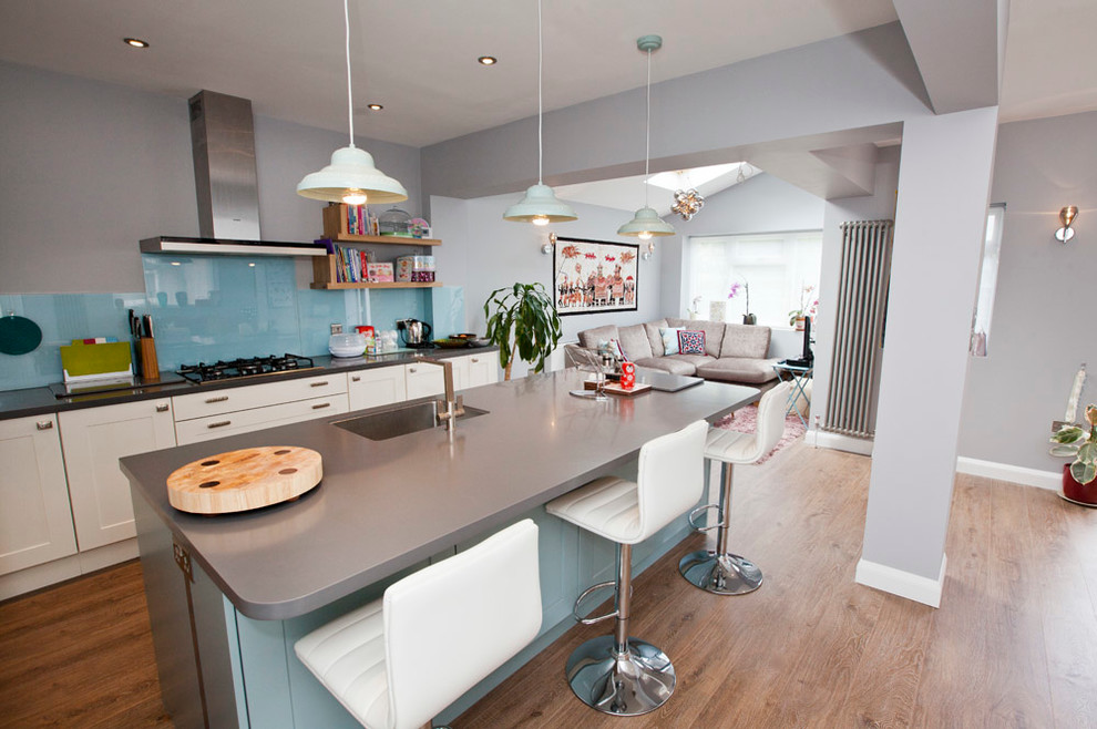 Medium sized eclectic l-shaped open plan kitchen in Other with a built-in sink, recessed-panel cabinets, white cabinets, composite countertops, blue splashback, glass sheet splashback, stainless steel appliances, medium hardwood flooring and an island.