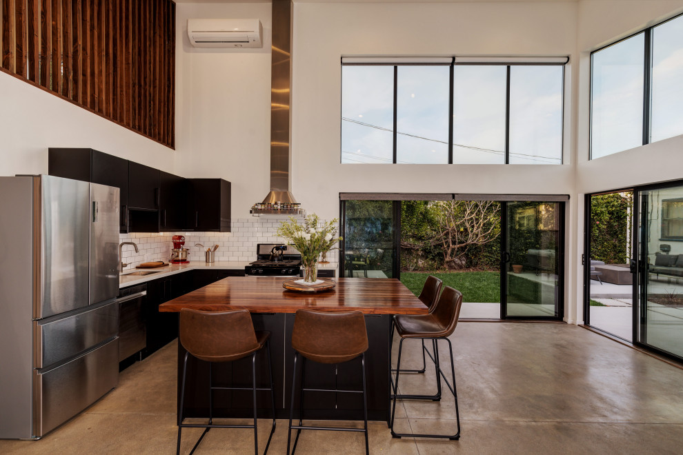 Example of a small trendy l-shaped concrete floor and gray floor open concept kitchen design in Los Angeles with a single-bowl sink, flat-panel cabinets, black cabinets, wood countertops, white backsplash, ceramic backsplash, stainless steel appliances, an island and brown countertops