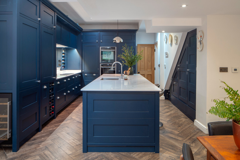 Design ideas for a traditional kitchen/diner in London with a submerged sink, shaker cabinets, blue cabinets, black appliances, an island, brown floors, white worktops and dark hardwood flooring.