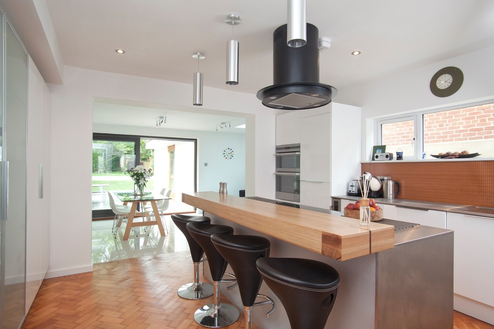 Medium sized contemporary galley kitchen/diner in Oxfordshire with flat-panel cabinets, white cabinets, stainless steel worktops, stainless steel appliances, medium hardwood flooring, an island, brown floors and brown splashback.