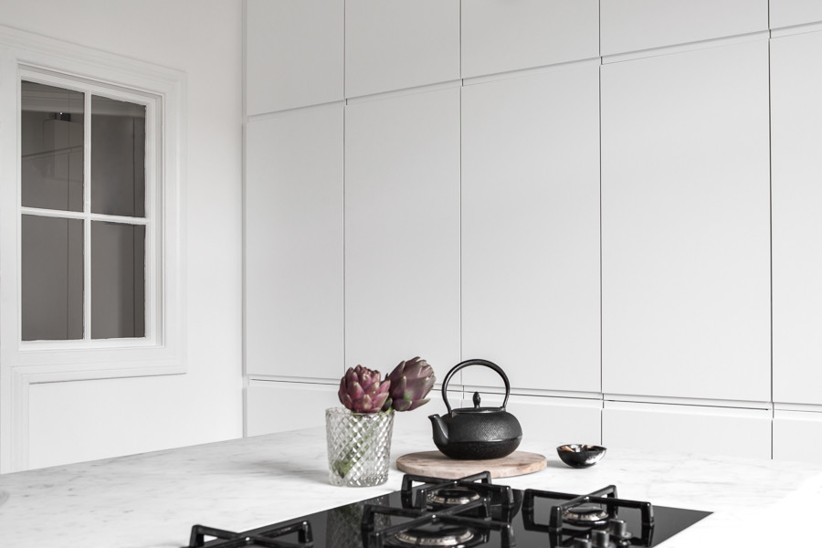 Minimalistisk inredning av ett stort, avskilt vit vitt kök, med släta luckor, vita skåp, marmorbänkskiva, vitt stänkskydd, stänkskydd i marmor, svarta vitvaror, ljust trägolv, en köksö och en integrerad diskho