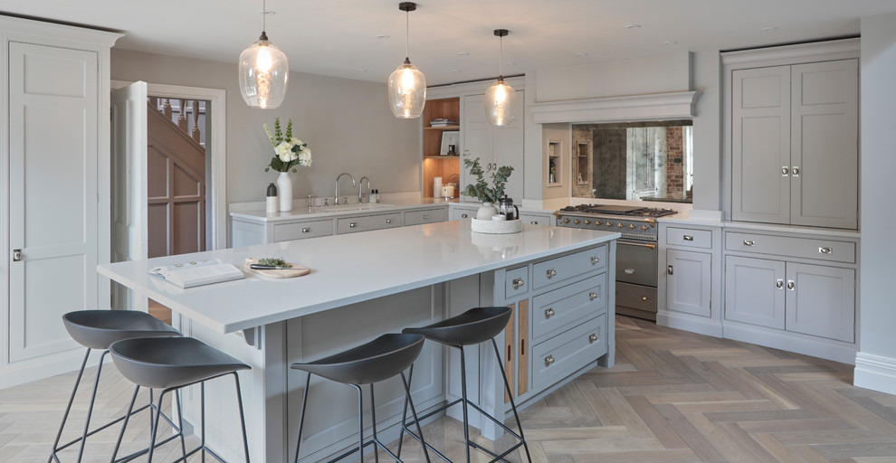 Example of a transitional light wood floor and brown floor kitchen design in Hertfordshire with shaker cabinets, gray cabinets, mirror backsplash, stainless steel appliances, an island and white countertops