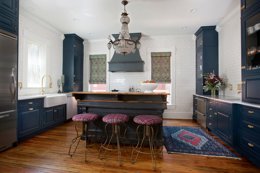 Example of an ornate galley medium tone wood floor and brown floor kitchen design in Denver with a farmhouse sink, raised-panel cabinets, blue cabinets, white backsplash, subway tile backsplash, stainless steel appliances, an island and white countertops