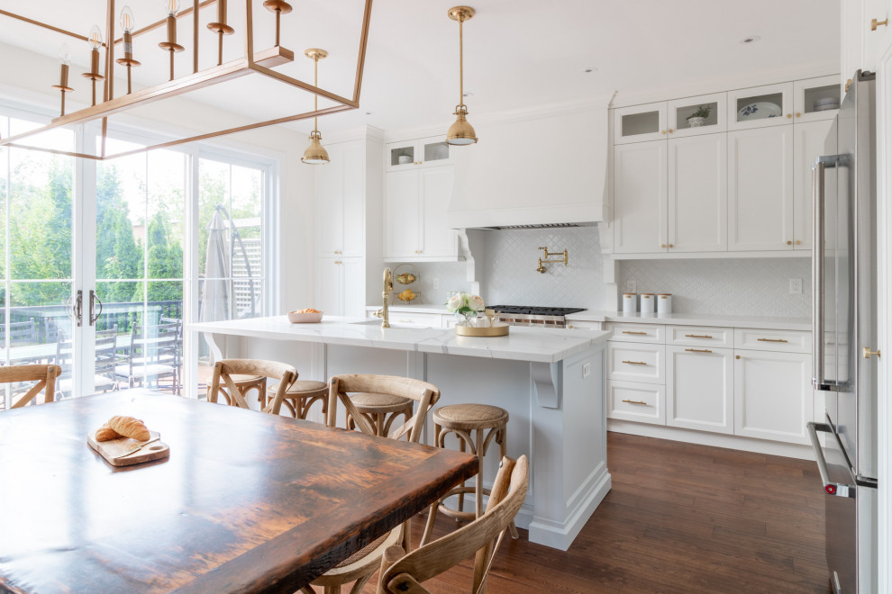 Photo of a large traditional galley kitchen/diner in Toronto with a belfast sink, shaker cabinets, white cabinets, engineered stone countertops, white splashback, porcelain splashback, stainless steel appliances, medium hardwood flooring, an island, brown floors and white worktops.
