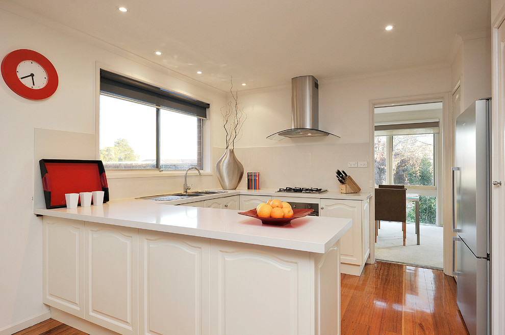 Small minimalist u-shaped medium tone wood floor eat-in kitchen photo in Melbourne with a double-bowl sink, beaded inset cabinets, white cabinets, solid surface countertops, white backsplash, glass sheet backsplash, stainless steel appliances and an island
