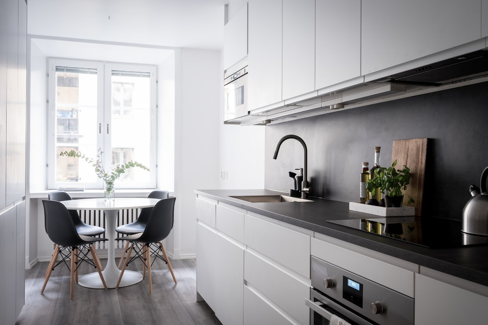 Diseño de cocina comedor lineal escandinava con armarios con paneles lisos, puertas de armario blancas, salpicadero negro, electrodomésticos de acero inoxidable, suelo de madera oscura, suelo gris y encimeras negras