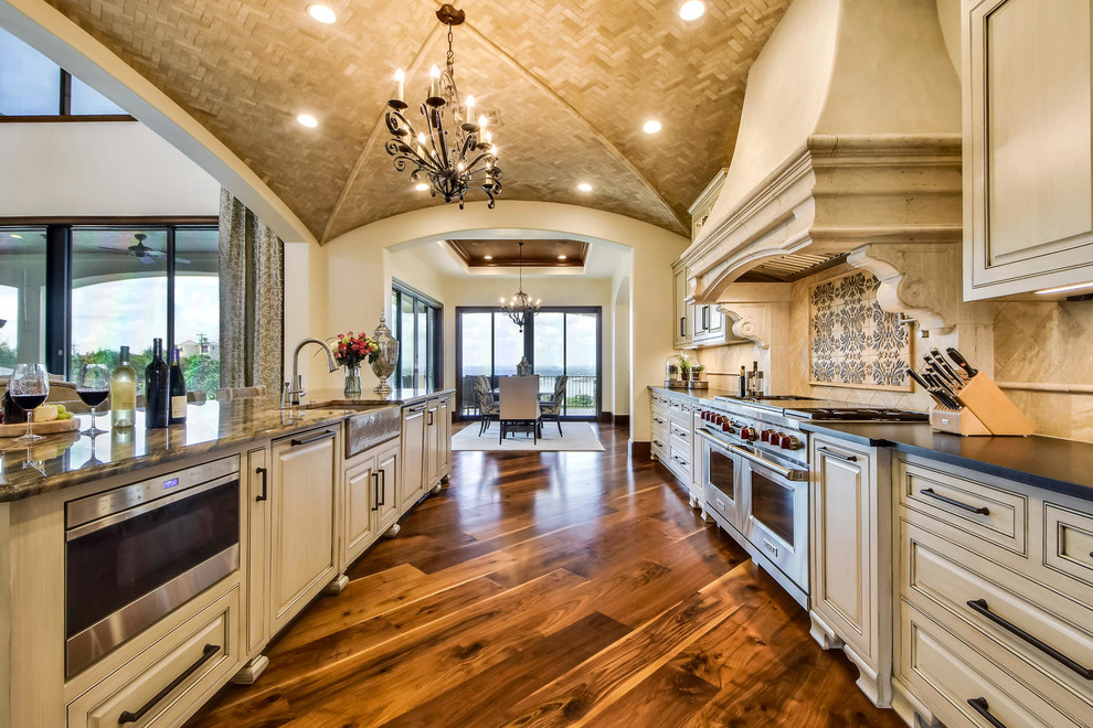 Example of a huge tuscan l-shaped medium tone wood floor open concept kitchen design in Austin with a farmhouse sink, raised-panel cabinets, distressed cabinets, quartzite countertops, beige backsplash, stone tile backsplash, stainless steel appliances and an island