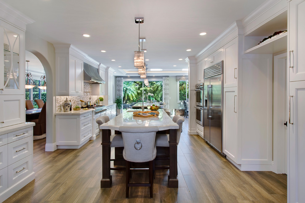 Design ideas for a large traditional galley kitchen/diner in Orange County with multiple islands, shaker cabinets, white cabinets, marble worktops, a submerged sink, beige splashback, mosaic tiled splashback, stainless steel appliances and medium hardwood flooring.