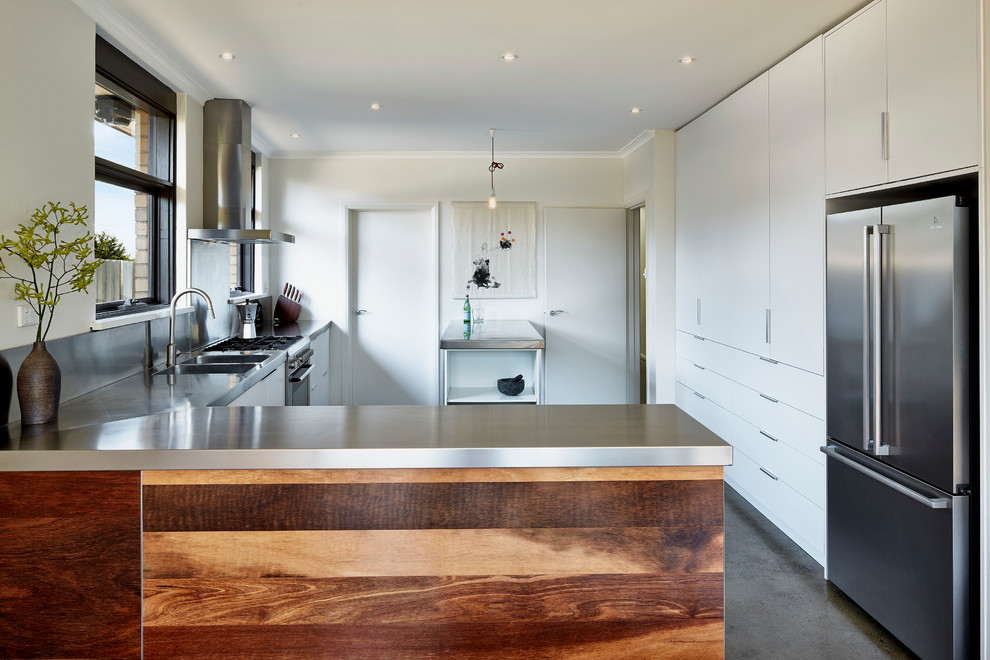 Idéer för att renovera ett mellanstort funkis l-kök, med en integrerad diskho, släta luckor, vita skåp, bänkskiva i rostfritt stål, rostfria vitvaror, betonggolv och en halv köksö