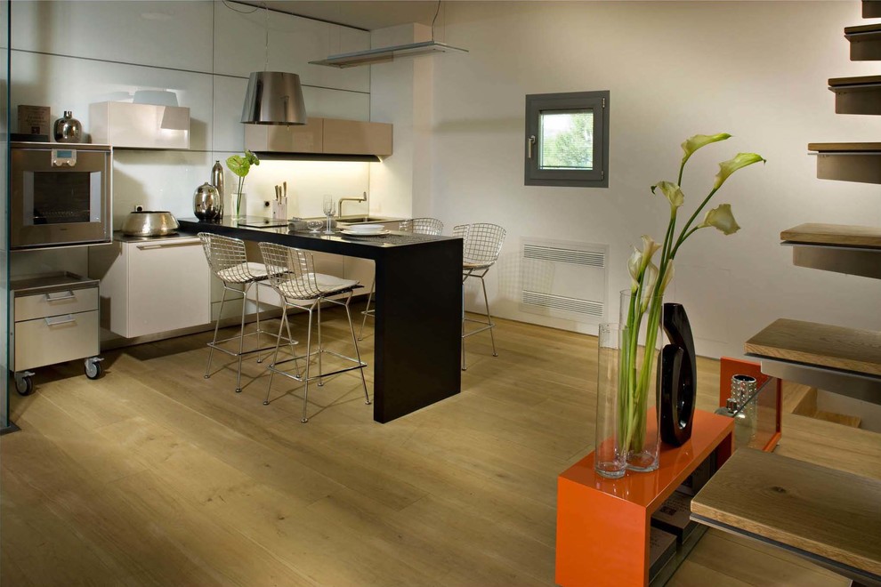 Foto på ett litet funkis linjärt kök och matrum, med en integrerad diskho, släta luckor, vita skåp, bänkskiva i rostfritt stål, vitt stänkskydd, rostfria vitvaror och ljust trägolv