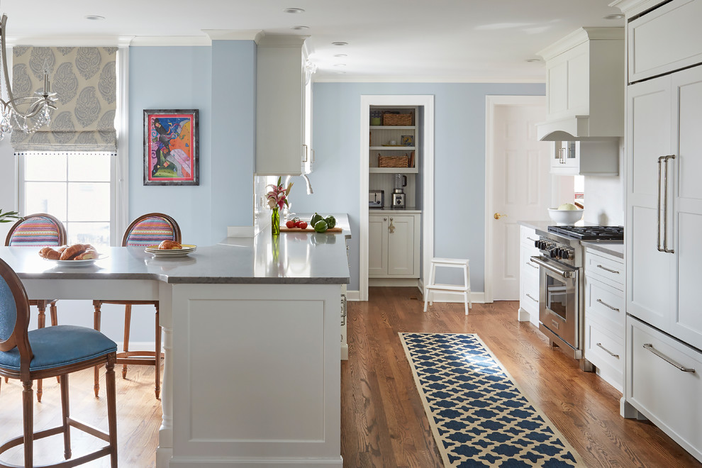 Idées déco pour une cuisine américaine classique avec un placard avec porte à panneau encastré, des portes de placard blanches, une crédence blanche, un électroménager en acier inoxydable, un sol en bois brun et un sol marron.