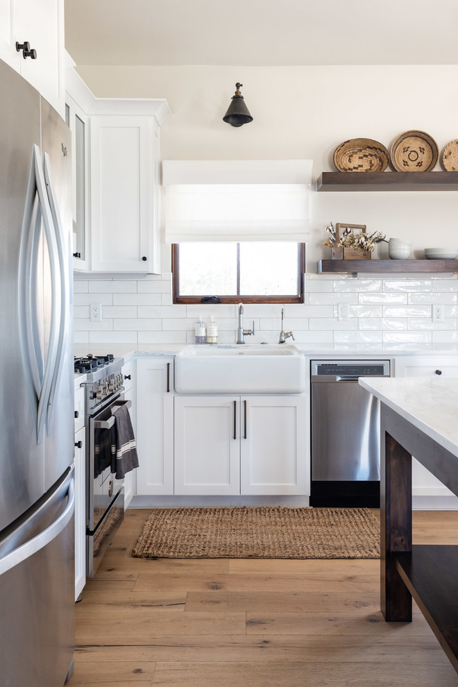 Example of a mid-sized cottage l-shaped light wood floor and brown floor open concept kitchen design in San Luis Obispo with a farmhouse sink, shaker cabinets, white cabinets, marble countertops, white backsplash, subway tile backsplash, stainless steel appliances, an island and white countertops