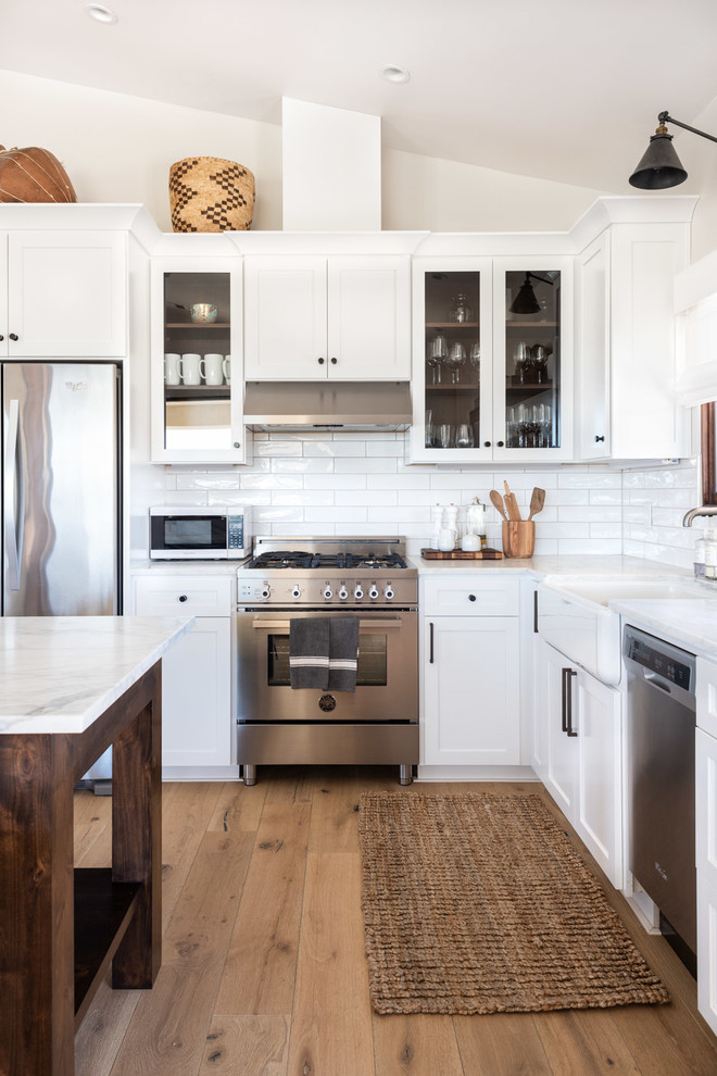 Example of a mid-sized country l-shaped light wood floor and brown floor open concept kitchen design in San Luis Obispo with a farmhouse sink, shaker cabinets, white cabinets, marble countertops, white backsplash, subway tile backsplash, stainless steel appliances, an island and white countertops