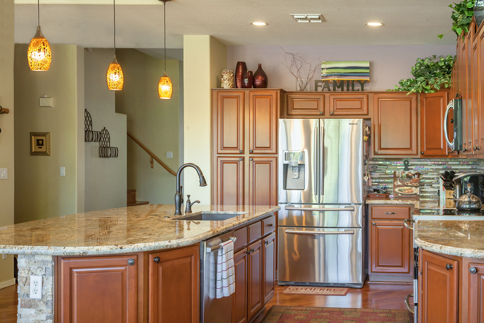 Inredning av ett klassiskt mellanstort l-kök, med en undermonterad diskho, luckor med infälld panel, bruna skåp, granitbänkskiva, flerfärgad stänkskydd, stänkskydd i glaskakel, rostfria vitvaror, laminatgolv, en köksö och brunt golv