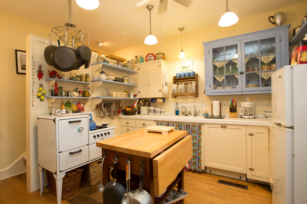 Bild på ett avskilt, litet shabby chic-inspirerat l-kök, med en undermonterad diskho, skåp i shakerstil, vita skåp, kaklad bänkskiva, vitt stänkskydd, stänkskydd i trä, vita vitvaror, ljust trägolv, en köksö och brunt golv