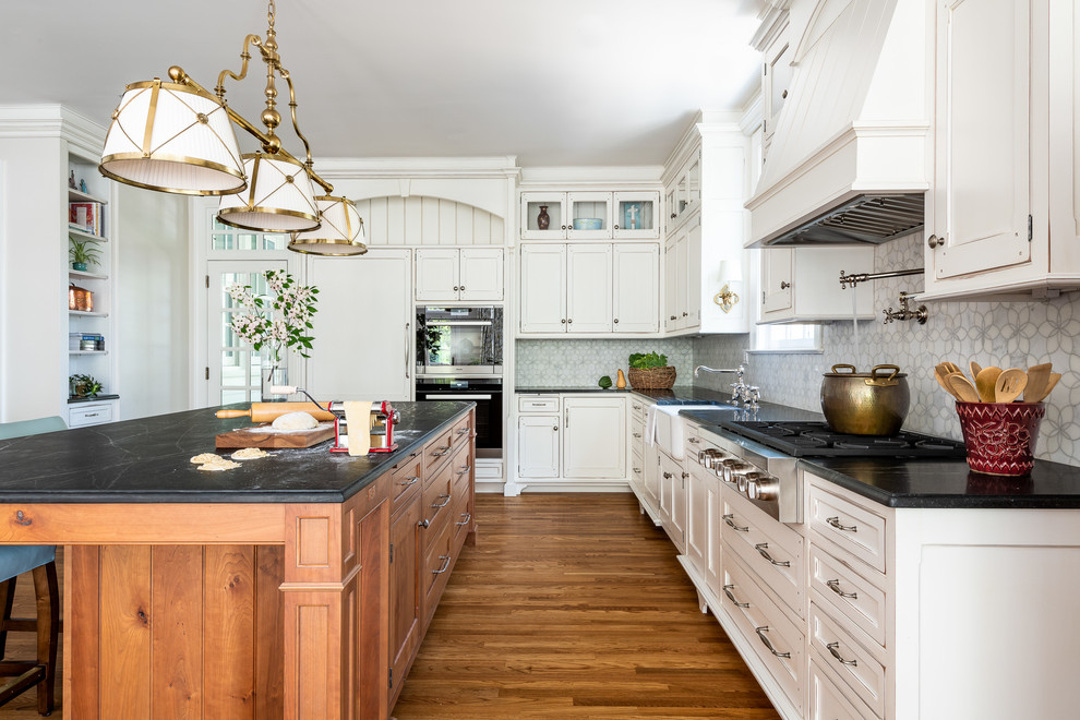 Inspiration for a large transitional u-shaped medium tone wood floor and brown floor enclosed kitchen remodel in DC Metro with a farmhouse sink, recessed-panel cabinets, white cabinets, soapstone countertops, white backsplash, paneled appliances, an island and black countertops