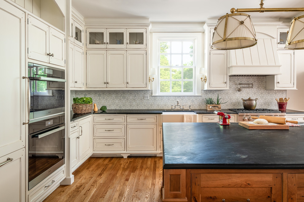 Large classic u-shaped enclosed kitchen in DC Metro with a belfast sink, recessed-panel cabinets, white cabinets, soapstone worktops, white splashback, integrated appliances, medium hardwood flooring, an island, brown floors and black worktops.