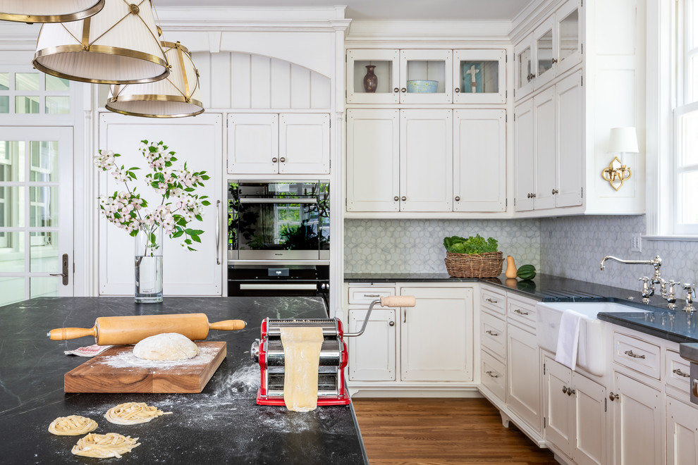 Example of a large transitional u-shaped medium tone wood floor and brown floor enclosed kitchen design in DC Metro with a farmhouse sink, recessed-panel cabinets, white cabinets, soapstone countertops, white backsplash, paneled appliances, an island and black countertops