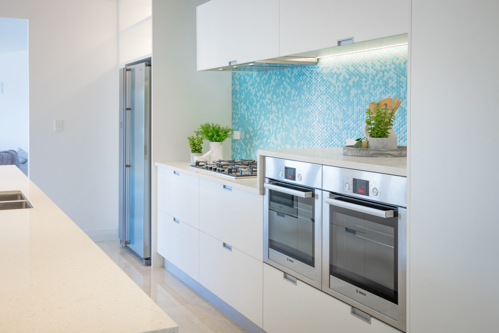 Maritim inredning av ett mellanstort linjärt kök med öppen planlösning, med en undermonterad diskho, släta luckor, vita skåp, granitbänkskiva, grönt stänkskydd, stänkskydd i mosaik, rostfria vitvaror, marmorgolv, en köksö och vitt golv
