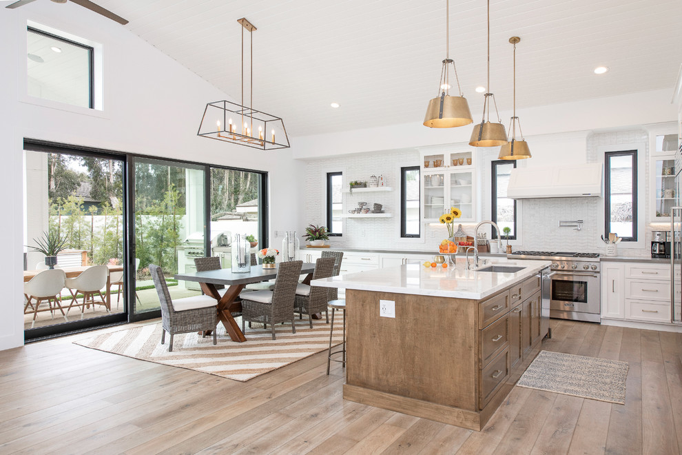 Inspiration for a large farmhouse l-shaped kitchen/diner in Orange County with a submerged sink, shaker cabinets, white cabinets, quartz worktops, white splashback, metro tiled splashback, stainless steel appliances, medium hardwood flooring, an island, brown floors and white worktops.