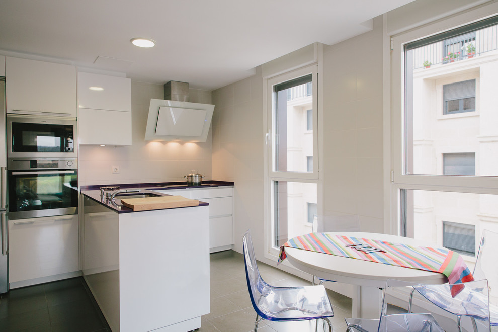 Источник вдохновения для домашнего уюта: угловая кухня-гостиная среднего размера в современном стиле с одинарной мойкой, плоскими фасадами, белыми фасадами, техникой из нержавеющей стали, полом из керамической плитки и полуостровом