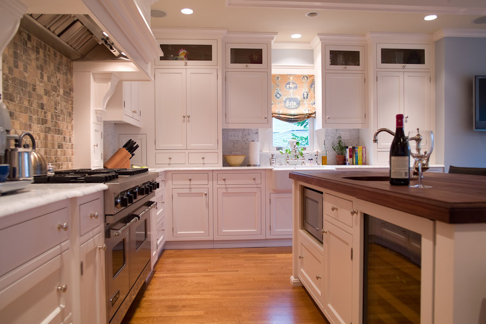 Example of a mid-sized classic l-shaped light wood floor enclosed kitchen design in Seattle with a farmhouse sink, shaker cabinets, white cabinets, marble countertops, beige backsplash, stone tile backsplash, stainless steel appliances and an island