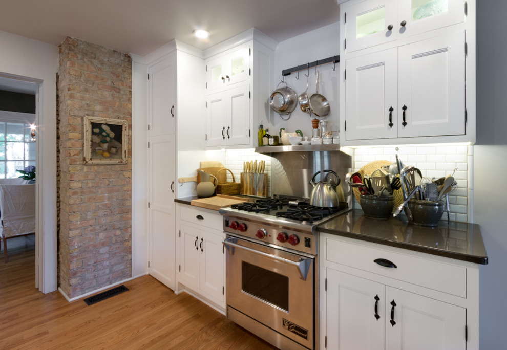 Photo of a large country u-shaped kitchen pantry in Chicago with a belfast sink, light wood cabinets, engineered stone countertops, white splashback, metro tiled splashback, stainless steel appliances, light hardwood flooring, an island and shaker cabinets.