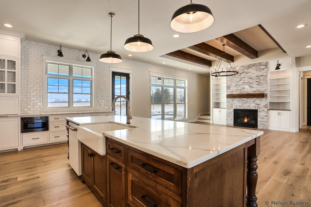 Photo of a medium sized farmhouse l-shaped open plan kitchen in Chicago with a belfast sink, shaker cabinets, white cabinets, engineered stone countertops, white splashback, metro tiled splashback, white appliances, light hardwood flooring, an island, brown floors, white worktops and exposed beams.