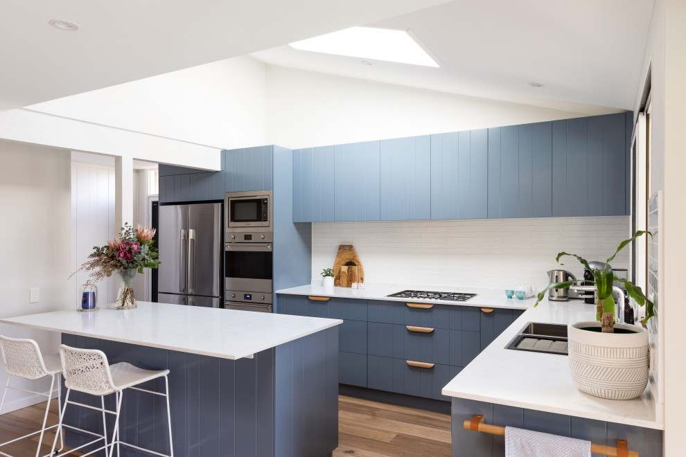 Foto på ett mellanstort funkis vit kök, med blå skåp, bänkskiva i kvarts, vitt stänkskydd, stänkskydd i porslinskakel, rostfria vitvaror, en dubbel diskho, mellanmörkt trägolv och en köksö