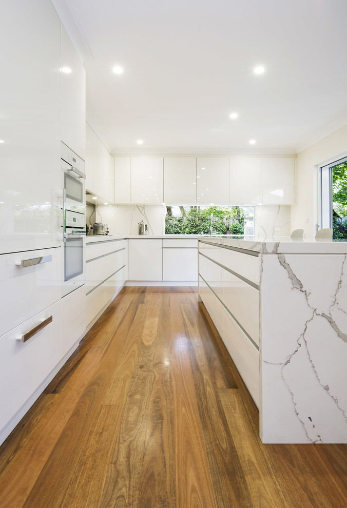 Exempel på ett mellanstort modernt kök, med en undermonterad diskho, släta luckor, vita skåp, bänkskiva i kvarts, stänkskydd med metallisk yta, spegel som stänkskydd, vita vitvaror, mellanmörkt trägolv och en köksö