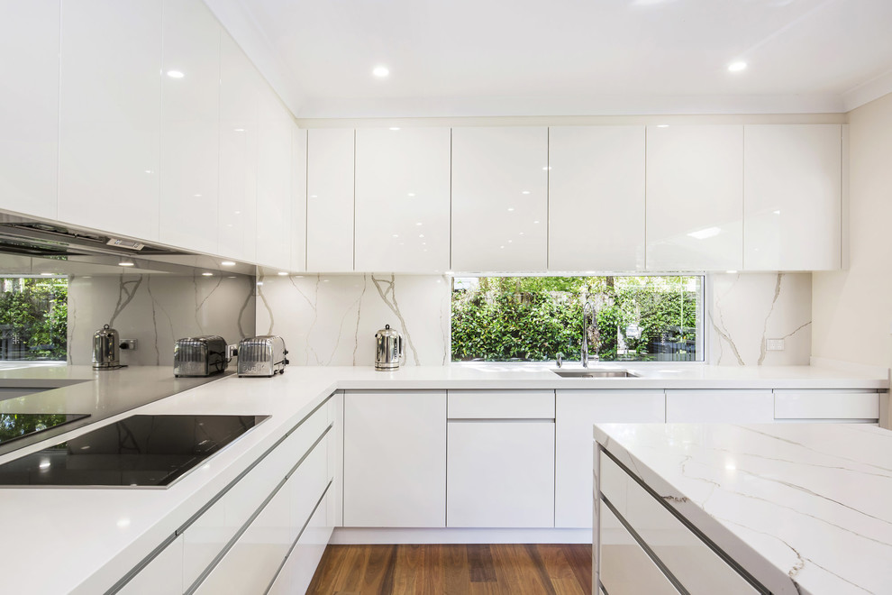 Bild på ett mellanstort funkis kök, med en undermonterad diskho, släta luckor, vita skåp, bänkskiva i kvarts, stänkskydd med metallisk yta, spegel som stänkskydd, vita vitvaror, mellanmörkt trägolv och en köksö