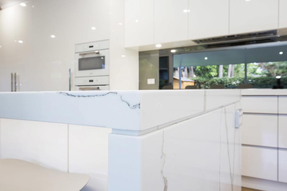 Modern inredning av ett mellanstort kök, med en undermonterad diskho, släta luckor, vita skåp, bänkskiva i kvarts, stänkskydd med metallisk yta, spegel som stänkskydd, vita vitvaror, mellanmörkt trägolv och en köksö