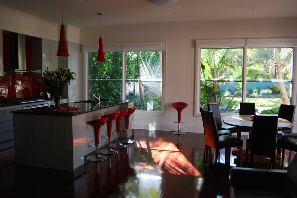 Foto på ett mellanstort funkis kök, med en undermonterad diskho, släta luckor, vita skåp, rött stänkskydd, glaspanel som stänkskydd, rostfria vitvaror, mörkt trägolv och en köksö