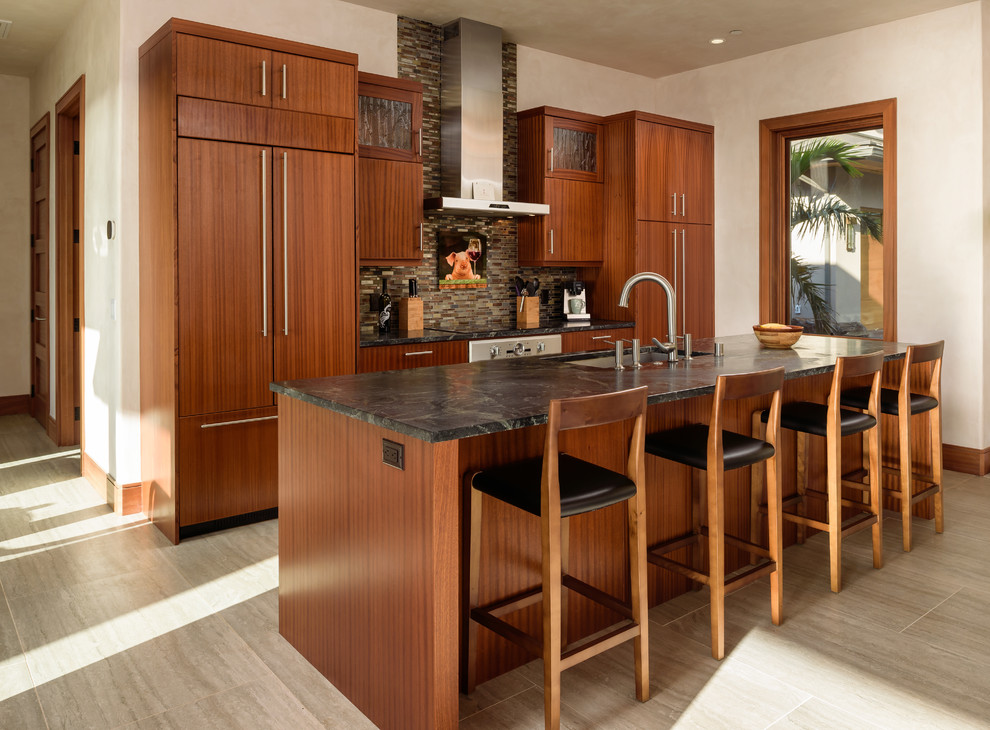 This is an example of a world-inspired galley kitchen in Hawaii with an island, a submerged sink, flat-panel cabinets, medium wood cabinets, brown splashback, matchstick tiled splashback, integrated appliances, beige floors and black worktops.
