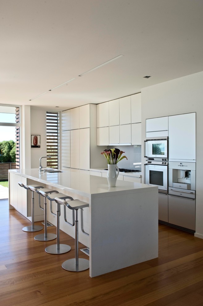 Inspiration for a contemporary kitchen/diner in Wellington with a double-bowl sink, white cabinets, engineered stone countertops, grey splashback, glass sheet splashback, stainless steel appliances, light hardwood flooring, an island and flat-panel cabinets.
