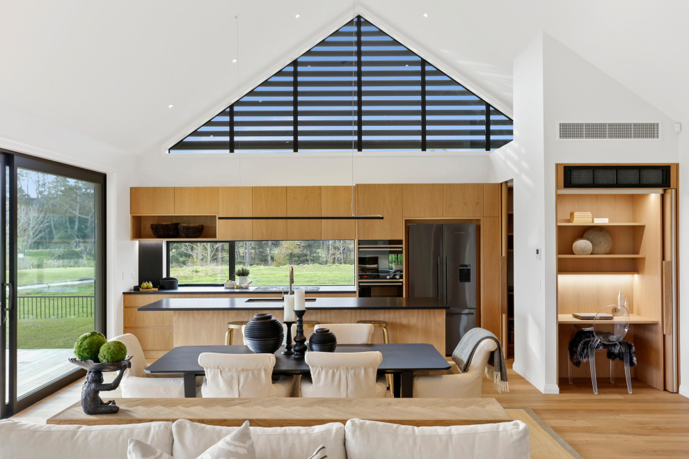 Photo of a contemporary galley open plan kitchen in Auckland with a submerged sink, flat-panel cabinets, medium wood cabinets, window splashback, stainless steel appliances, medium hardwood flooring, an island, brown floors, black worktops and a vaulted ceiling.