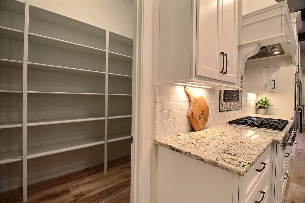Photo of an expansive classic l-shaped kitchen pantry in Portland with a submerged sink, recessed-panel cabinets, white cabinets, granite worktops, metallic splashback, glass tiled splashback, integrated appliances, light hardwood flooring, an island and beige floors.
