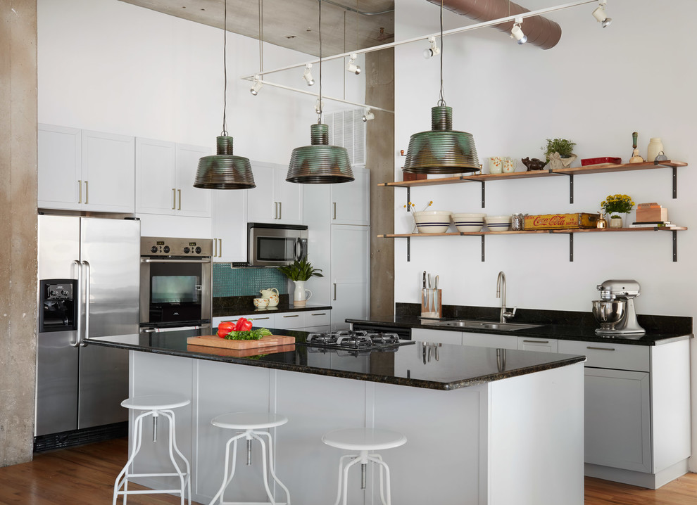 Photo of an urban kitchen in Chicago with a double-bowl sink, recessed-panel cabinets, grey cabinets, granite worktops, green splashback, stainless steel appliances, an island, black worktops, mosaic tiled splashback and medium hardwood flooring.