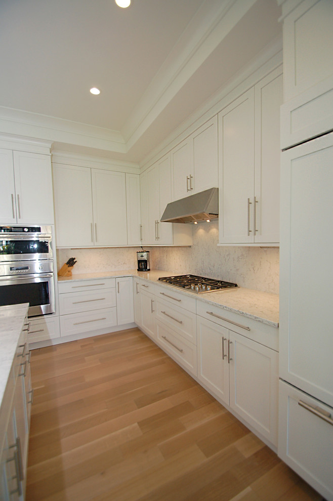 Example of a mid-sized transitional l-shaped light wood floor eat-in kitchen design in New York with an undermount sink, recessed-panel cabinets, white cabinets, quartz countertops, white backsplash, stone slab backsplash, paneled appliances and an island