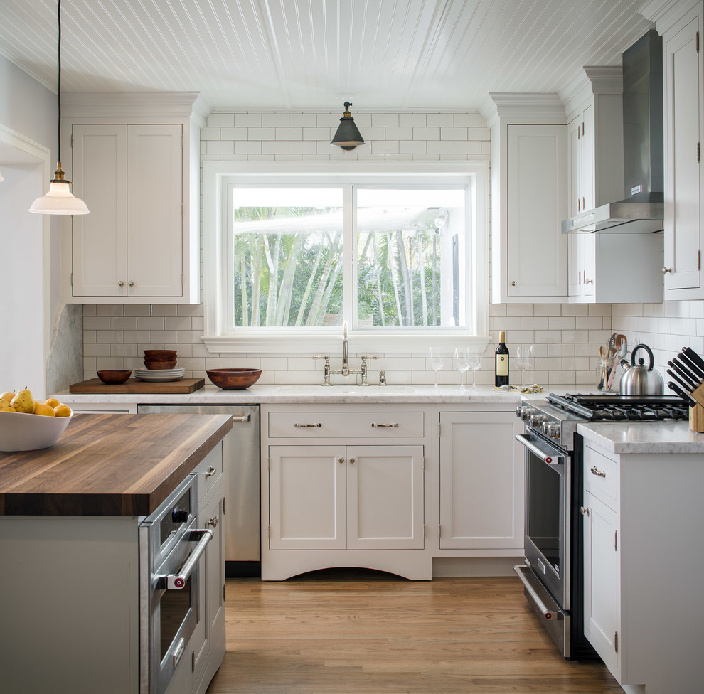 Bild på ett lantligt l-kök, med skåp i shakerstil, vita skåp, vitt stänkskydd, stänkskydd i tunnelbanekakel, rostfria vitvaror, mellanmörkt trägolv och en köksö