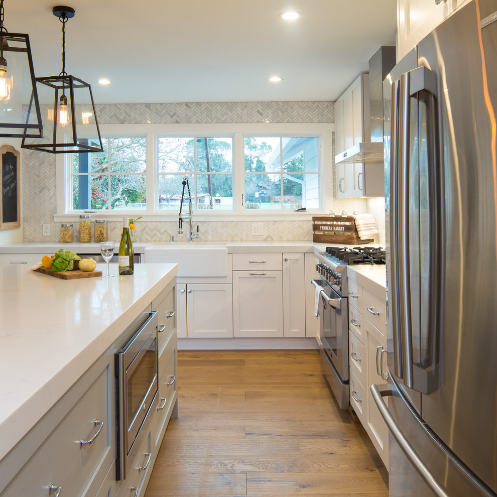 Medium sized farmhouse l-shaped open plan kitchen in San Francisco with a belfast sink, shaker cabinets, white cabinets, engineered stone countertops, white splashback, marble splashback, stainless steel appliances, medium hardwood flooring, an island and brown floors.