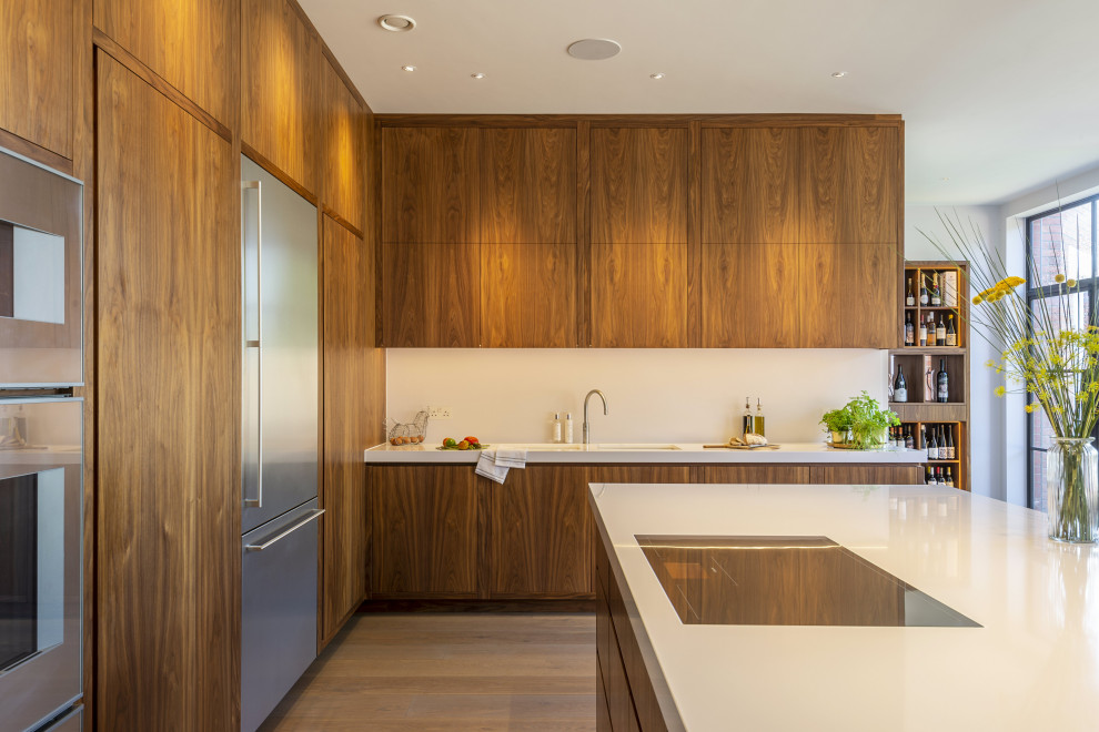 This is an example of a large midcentury l-shaped kitchen/diner in London with an integrated sink, flat-panel cabinets, medium wood cabinets, quartz worktops, white splashback, stone slab splashback, stainless steel appliances, medium hardwood flooring, an island, brown floors and white worktops.