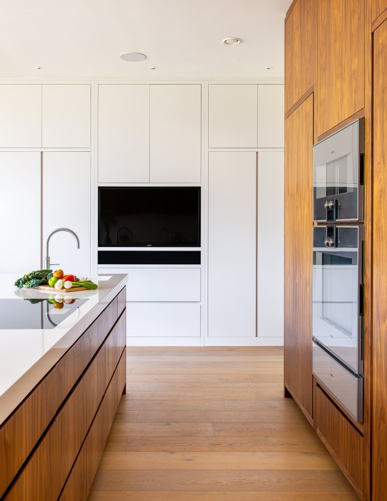 Foto på ett stort retro vit kök, med en integrerad diskho, släta luckor, skåp i mellenmörkt trä, bänkskiva i kvartsit, vitt stänkskydd, stänkskydd i sten, rostfria vitvaror, mellanmörkt trägolv, en köksö och brunt golv